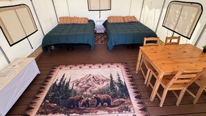 Een tent met twee bedden, een tafel, stoelen, een vloerkleed met een ontwerp van een beer, en een plafondventilator.