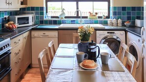 A kitchen with a dining table, chairs, a microwave, a sink, a washing machine, and a clock on the wall. - 3 bedroom accommodation in Rhostrehwfa Nr Llangefni (Rhostrehwfa Nr Llangefni)