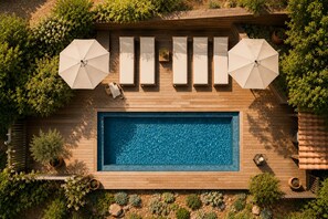 Una piscina rettangolare con un solarium in legno, circondata da vegetazione e due ombrelloni.