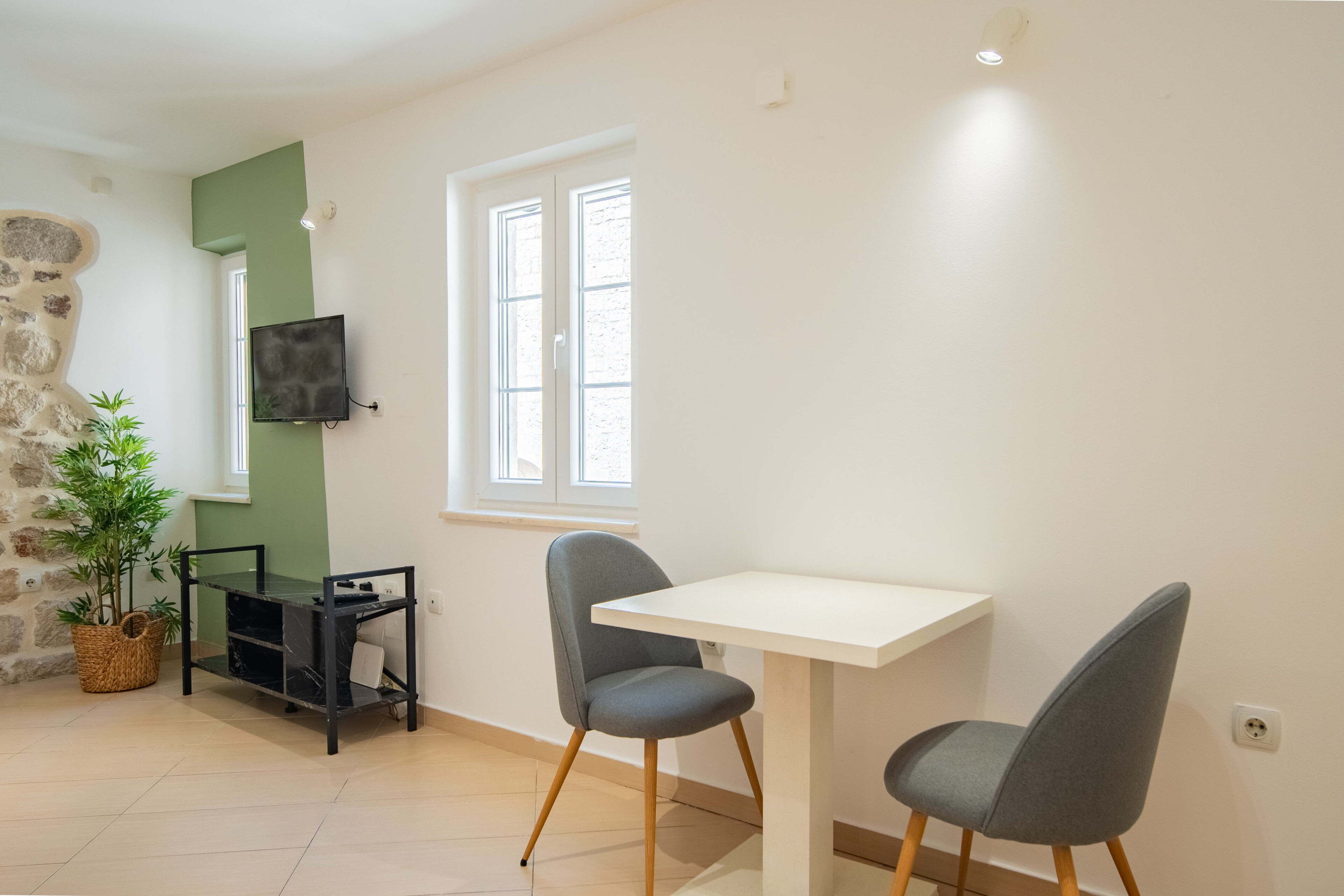 A room with a white table, two grey chairs, a TV on the wall, a stone wall, a window, and a plant.