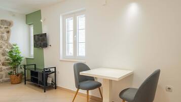 A room with a white table, two grey chairs, a TV on the wall, a stone wall, a window, and a plant.