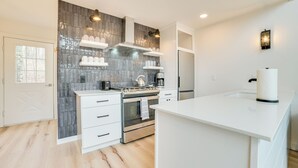 Una cocina moderna con armarios blancos, horno de acero inoxidable, estufa a gas y microondas integrado. Hay estantes blancos sobre la estufa y una encimera blanca.