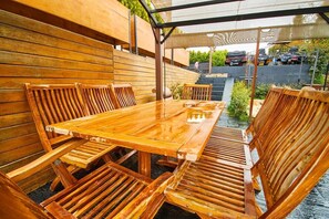 An outdoor wooden table and chairs set against a wooden wall, with a covered area overhead and a view of parked vehicles in the background.