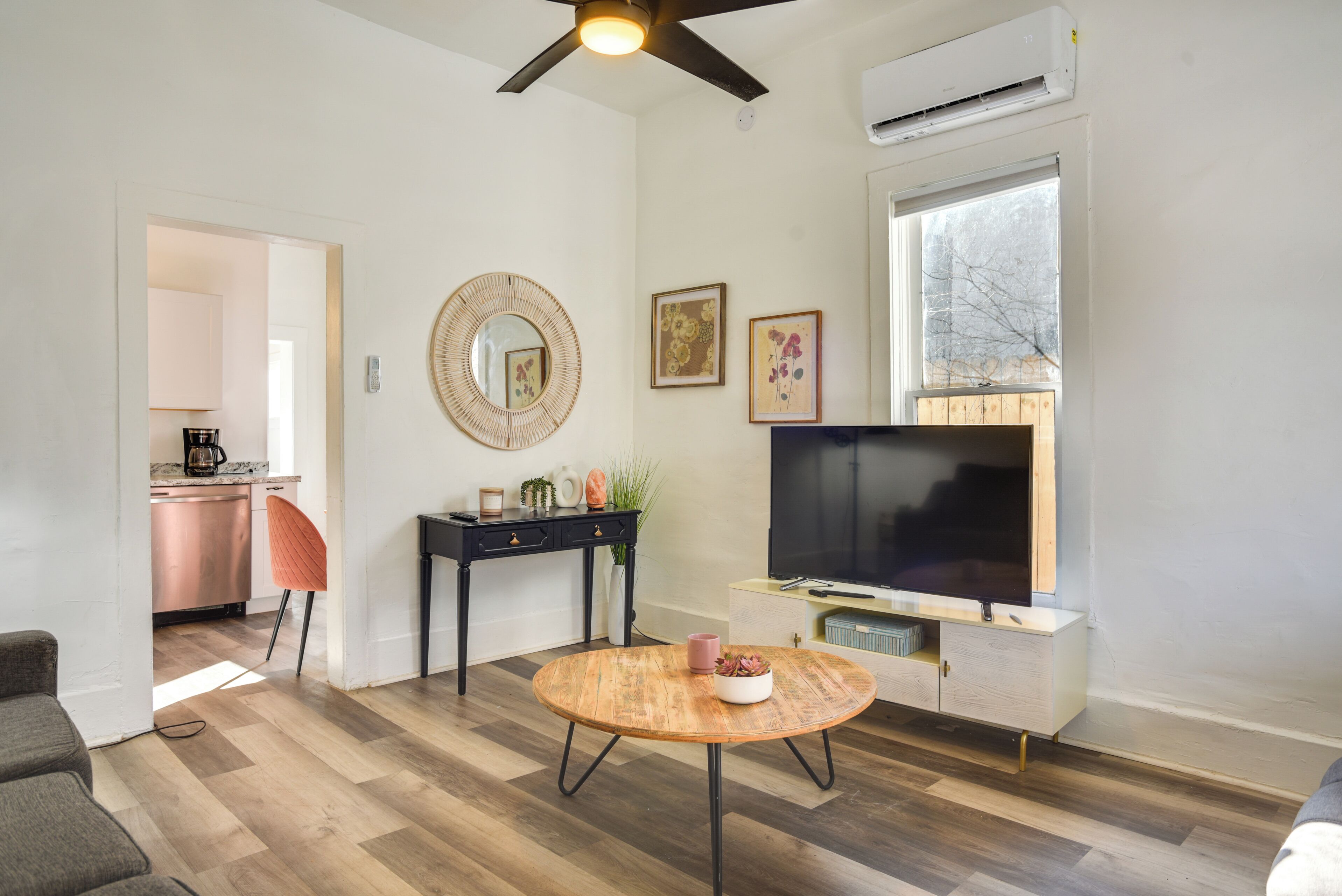 A modern living room with a wooden floor, a round coffee table, a TV on a white cabinet, a ceiling fan, and a kitchen in the background.