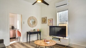 A modern living room with a wooden floor, a round coffee table, a TV on a white cabinet, a ceiling fan, and a kitchen in the background. - Near UNM & ABQ BioPark: East Dtwn Studio (Albuquerque)