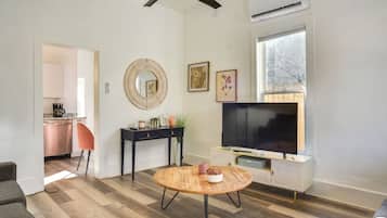 A modern living room with a wooden floor, a round coffee table, a TV on a white cabinet, a ceiling fan, and a kitchen in the background.
