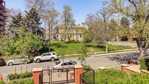 Un parking clos, avec une entrée en briques, situé à côté d’une route où sont garées des voitures et où se trouve un grand arbre.
