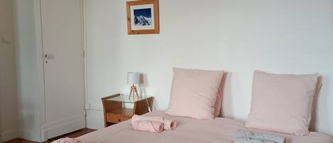A neatly made bed with pink bedding and decorative pillows, a small bedside table with a lamp, and a framed picture on the wall.