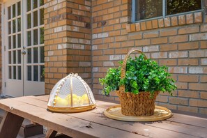 A wooden table with a basket of green plants and a yellow fruit basket with a protective mesh cover.