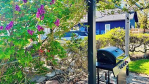 A barbecue grill in a garden with purple flowers and a blue house in the background. - Coeur d' Lovely (Coeur d'Alene)