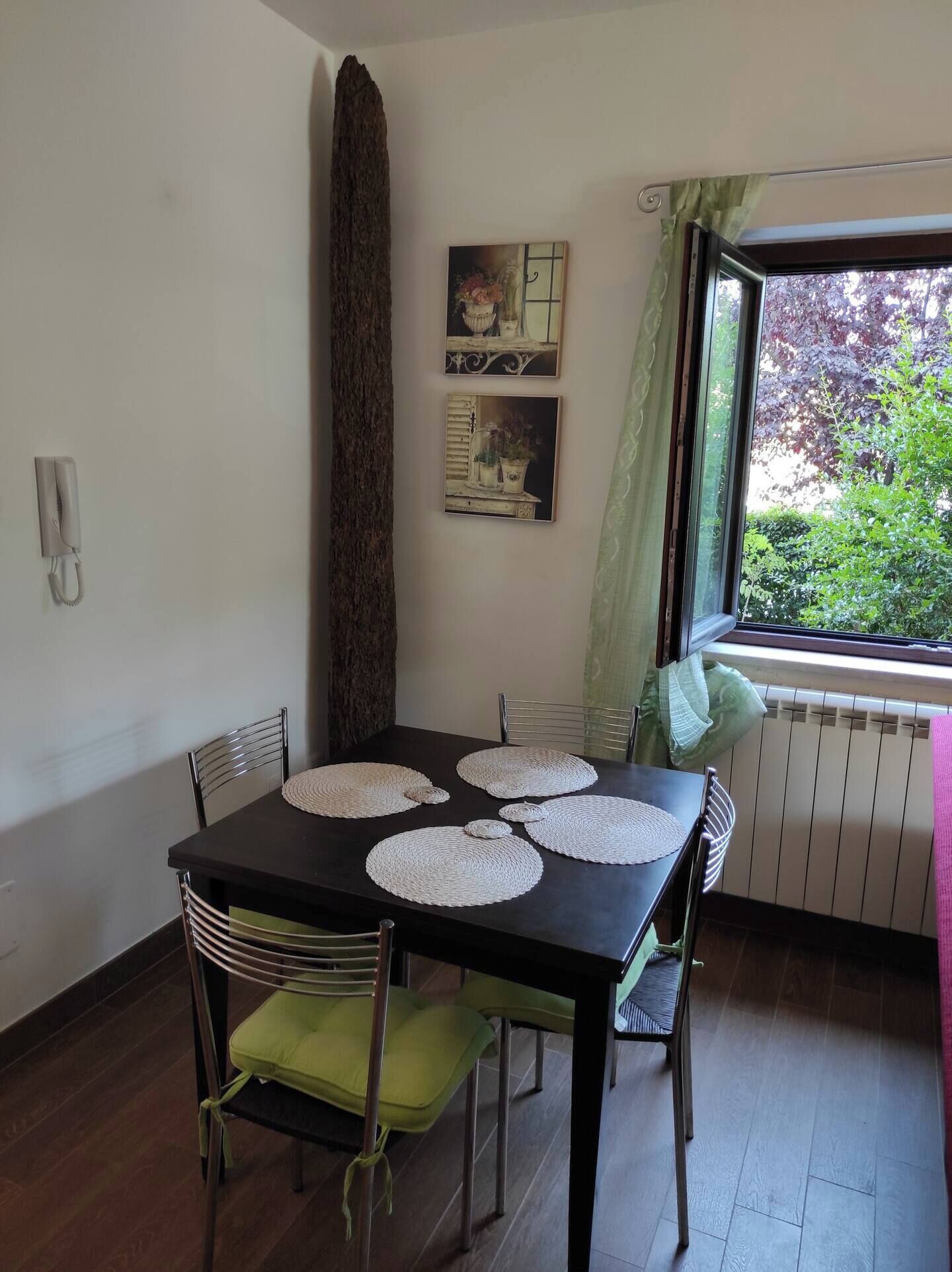A dining area with a dark wooden table, four chairs, and woven placemats. There are two framed pictures on the wall, a tall tree trunk, and a window with a view of greenery.