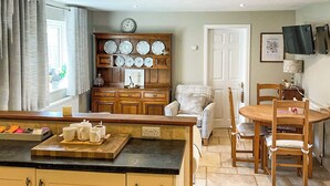 Une cuisine avec des meubles de rangement en bois, une table à manger avec des chaises, une horloge au mur et une fenêtre avec des rideaux.