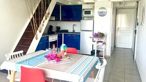 Eine kompakte Küche mit blauen Schränken, einer Mikrowelle und einem Kühlschrank. Im Vordergrund steht ein Esstisch mit Stühlen, und links ist eine Treppe zu sehen.