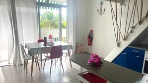 Ein Essbereich mit Tisch und Stühlen, eine Küchenzeile mit blauen Schränken und Blick auf Palmen durch eine Schiebetür.