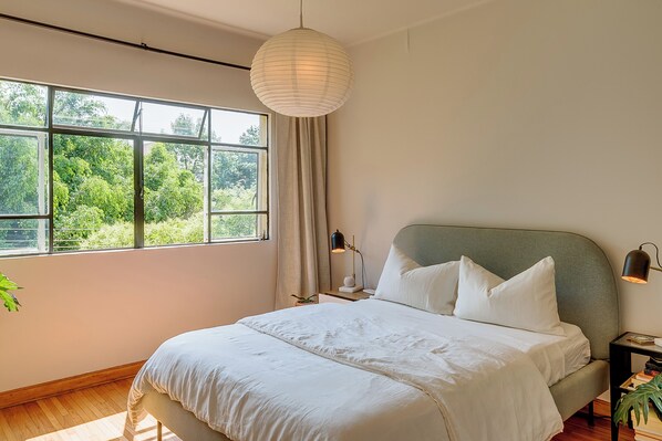 Ein Schlafzimmer mit einem großen Fenster, einem Bett mit weißer Bettwäsche, einem grünen Kopfteil und einer Pendelleuchte.