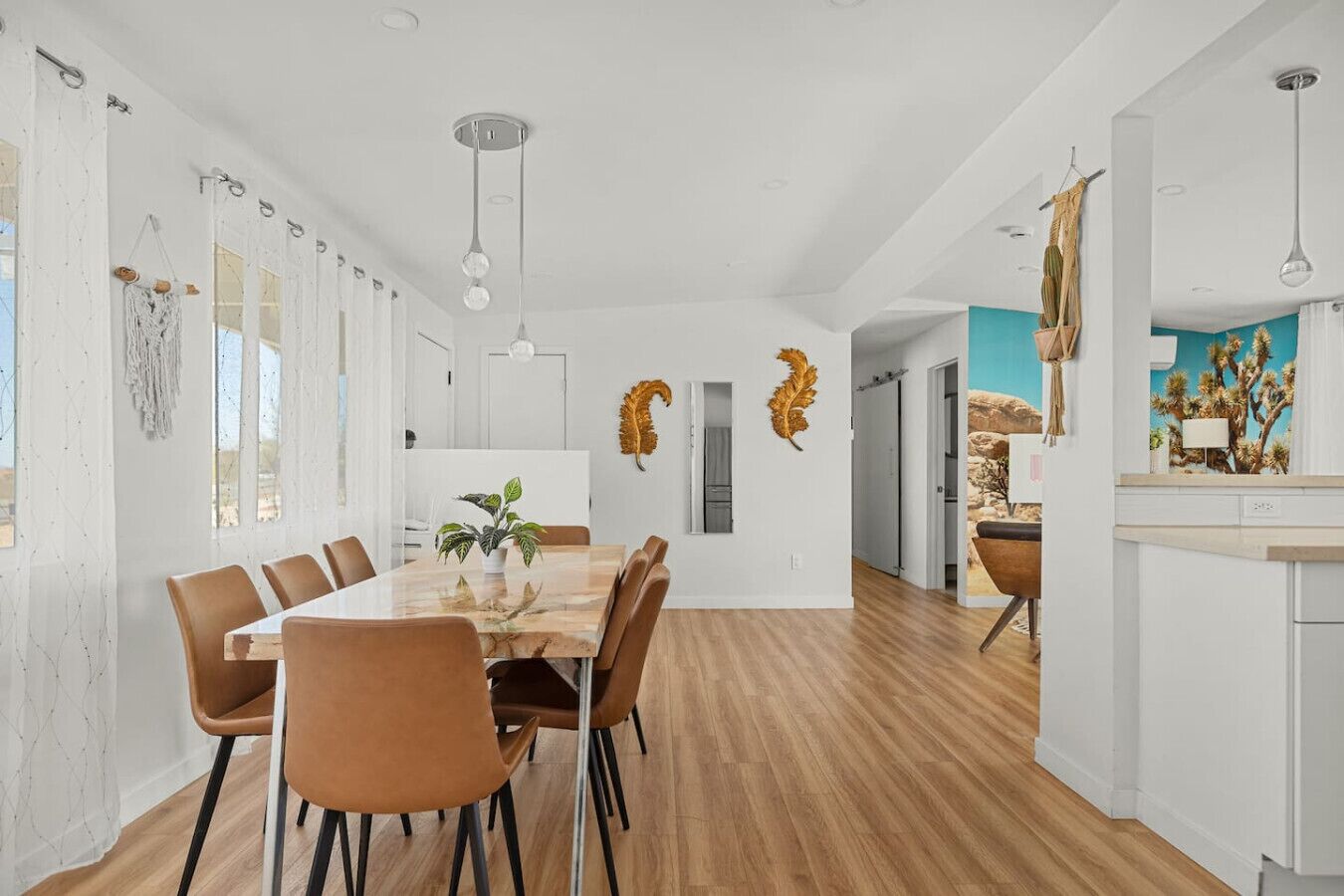 A modern dining room with a wooden table and chairs, a potted plant, and a kitchen visible through an open door.