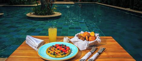 A dining table with a waffle, fruit, and a drink by a poolside.