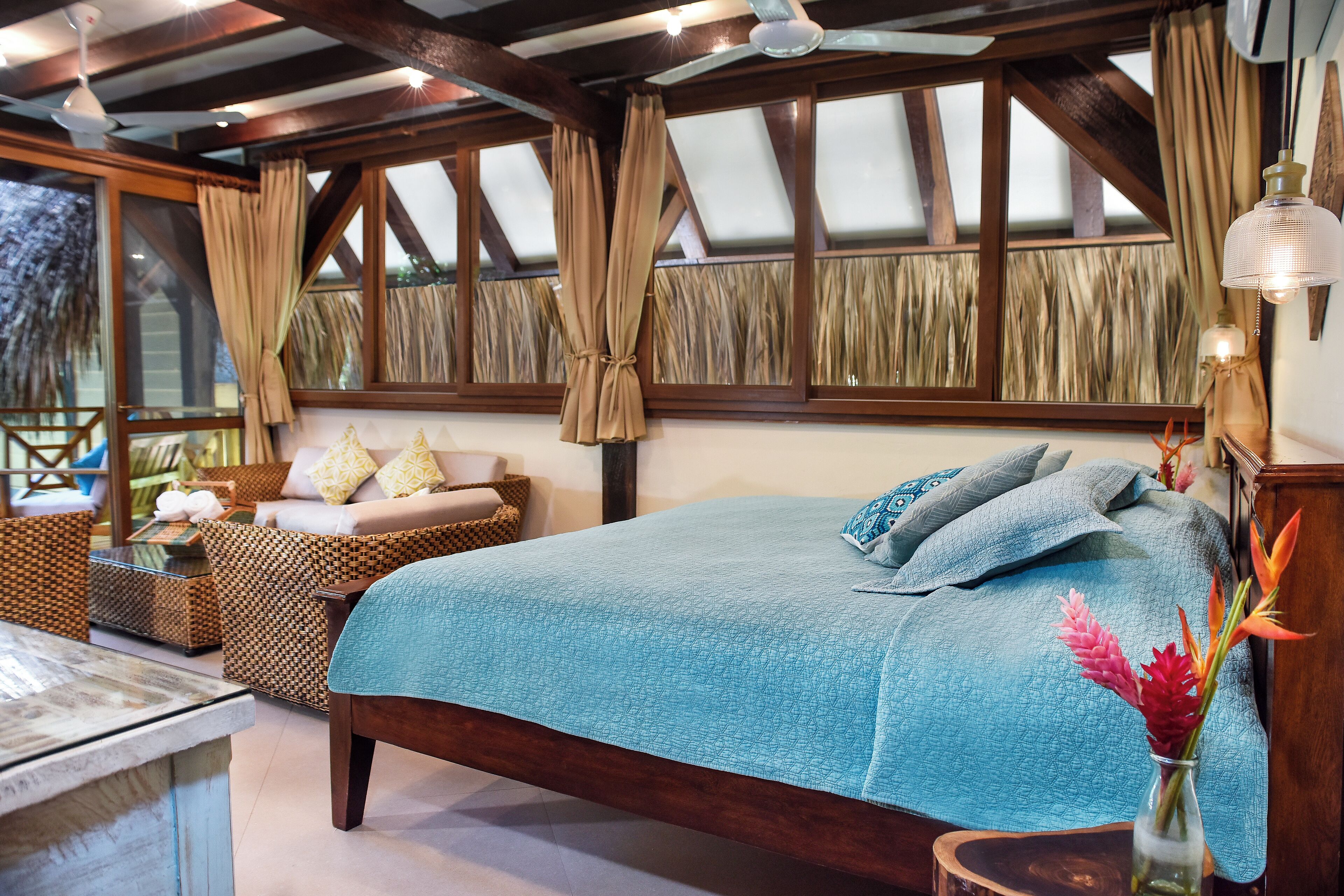 Quarto com uma cama grande, cabeceira de madeira, mesa de cabeceira e criadeira. Há uma janela com cortinas, uma luminária na parede e um vaso com flores na criadeira.