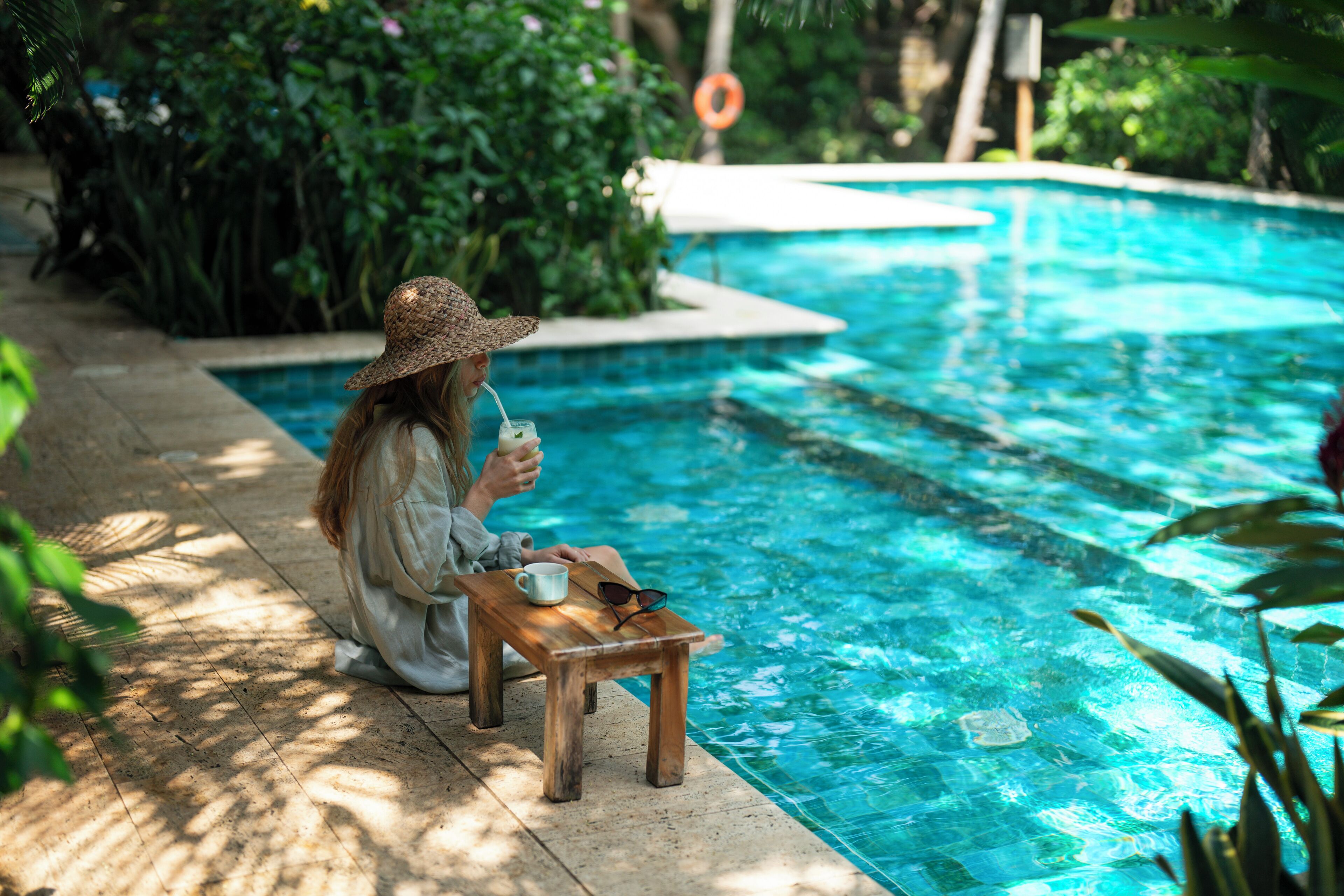 A person sitting by a pool with a drink, a cup, and sunglasses on a small table.