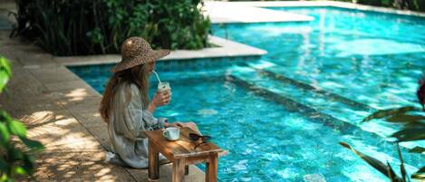 A person sitting by a pool with a drink, a cup, and sunglasses on a small table.