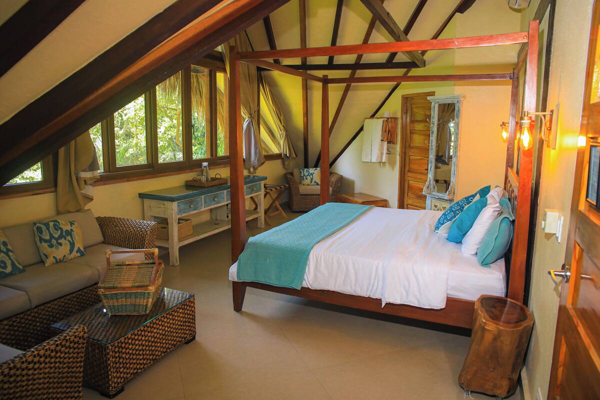 A bedroom with a wooden four-poster bed, a sofa, a small table, and a window with curtains.