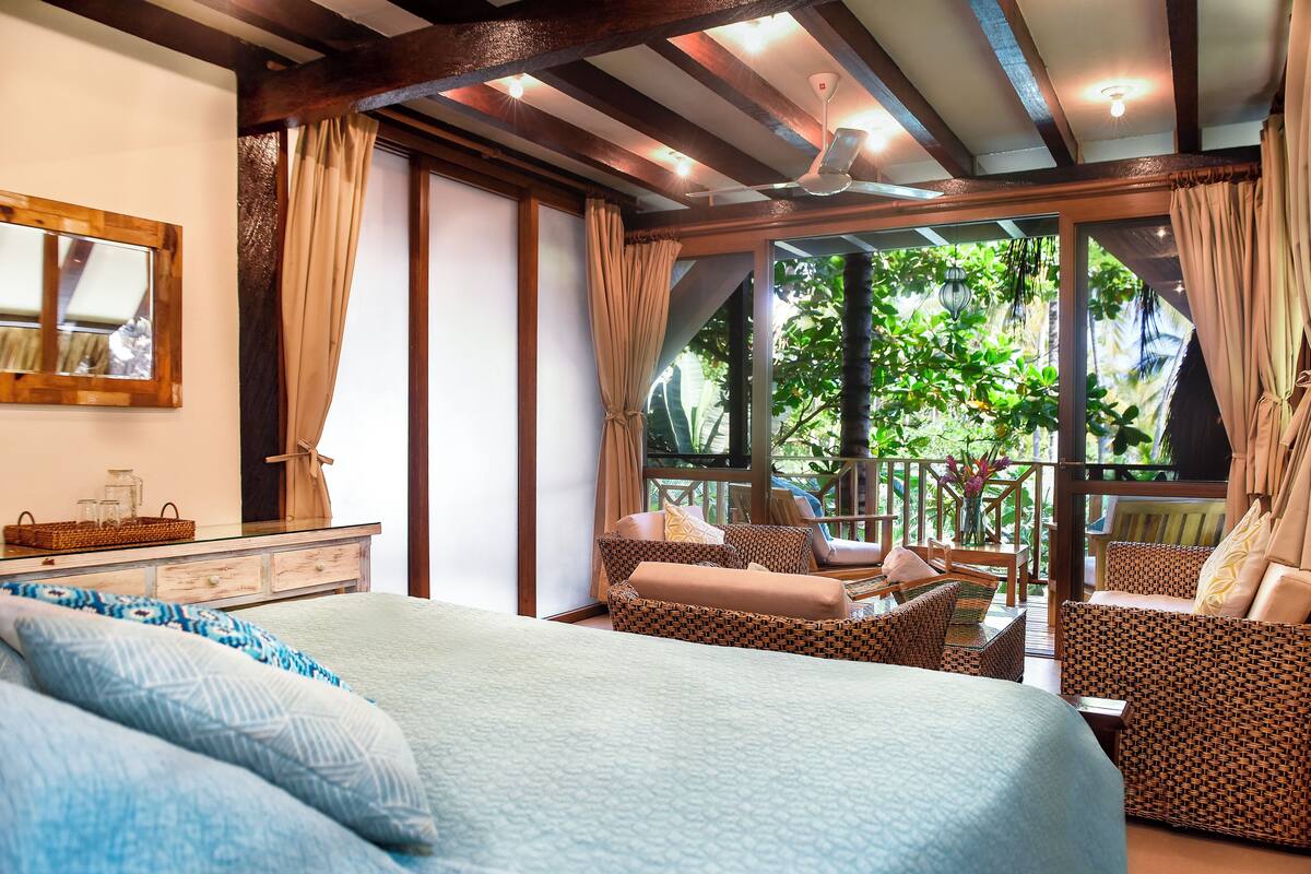 A spacious bedroom with a large bed, two wicker sofas, a wooden dresser, and a balcony with a view of lush greenery.