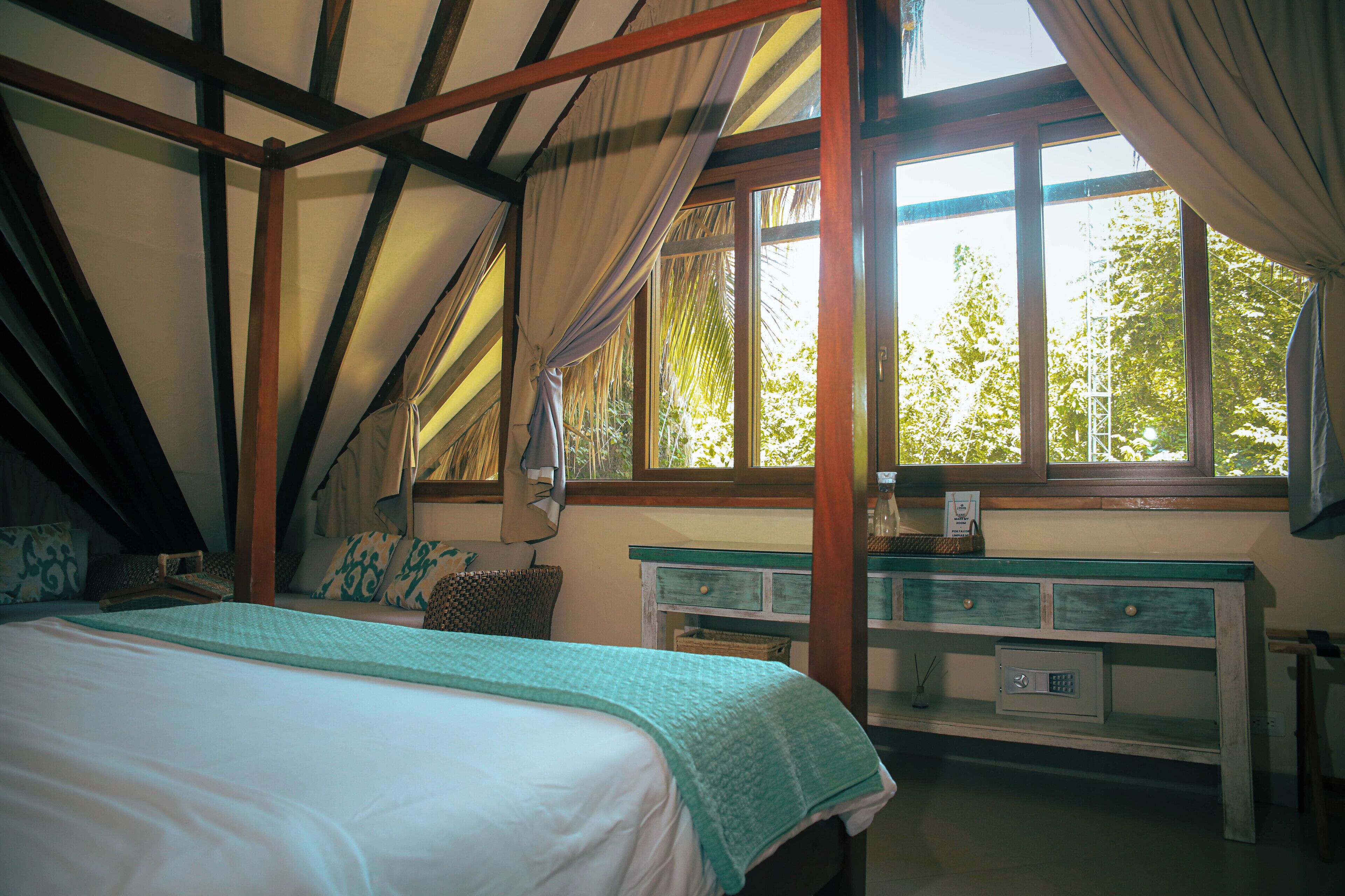 Une chambre à coucher avec un lit, une table de chevet, une fenêtre avec des rideaux et une vue sur les arbres à l’extérieur.