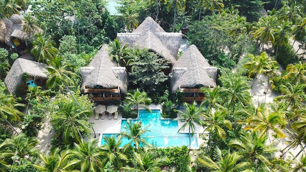 Een resort met bungalows met rieten daken, een centraal gelegen zwembad en weelderige tropische begroeiing.