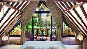 Een slaapkamer met een groot bed, gordijnen en uitzicht op een houten terras en weelderige begroeiing.