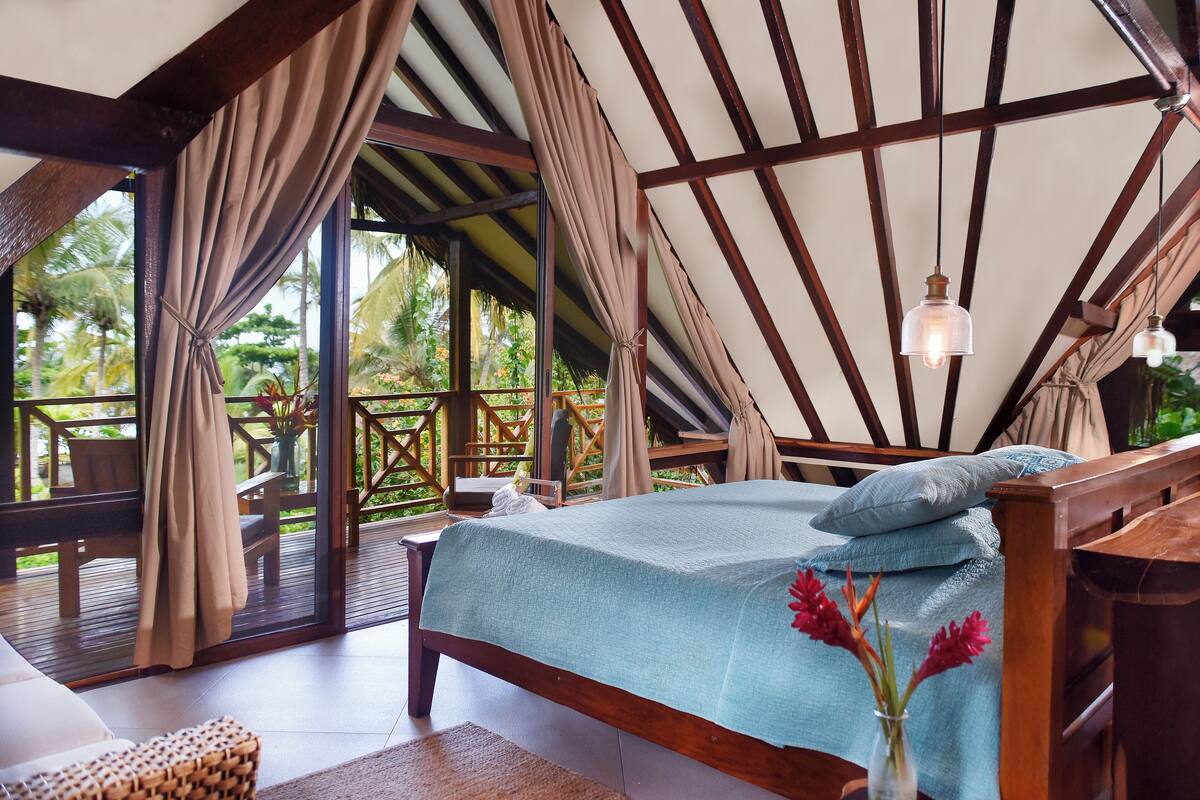 A bedroom with a bed, a wooden headboard, a nightstand, a vase with flowers, and a view of the outdoors.