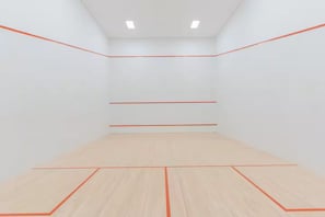 Quadra de squash com paredes brancas e piso de madeira demarcado com linhas vermelhas.