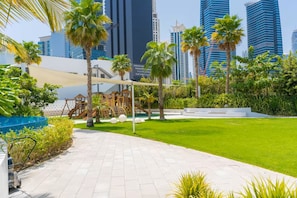 Un área exterior con jardines, un parque infantil, palmeras y una piscina.