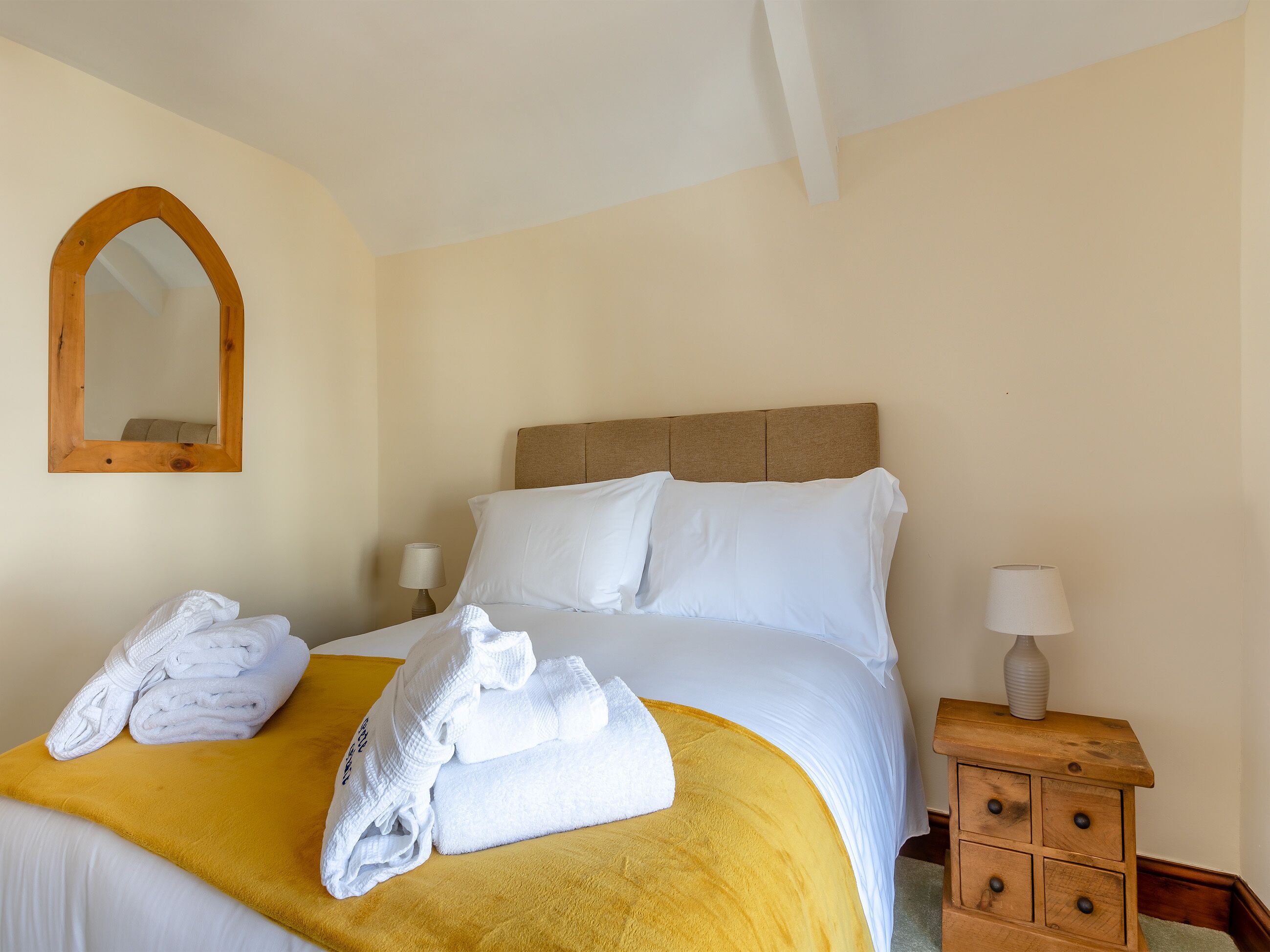 A neatly made bed with white linens and a yellow throw blanket, a wooden nightstand with a lamp, and a mirror on the wall.