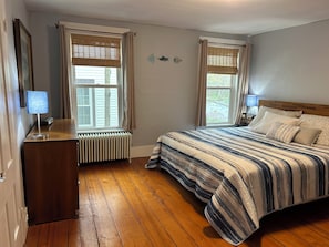 Ein Schlafzimmer mit Holzboden, einem Bett mit gestreifter Bettwäsche, einem Nachttisch mit Lampe und einem Heizkörper unter einem Fenster.