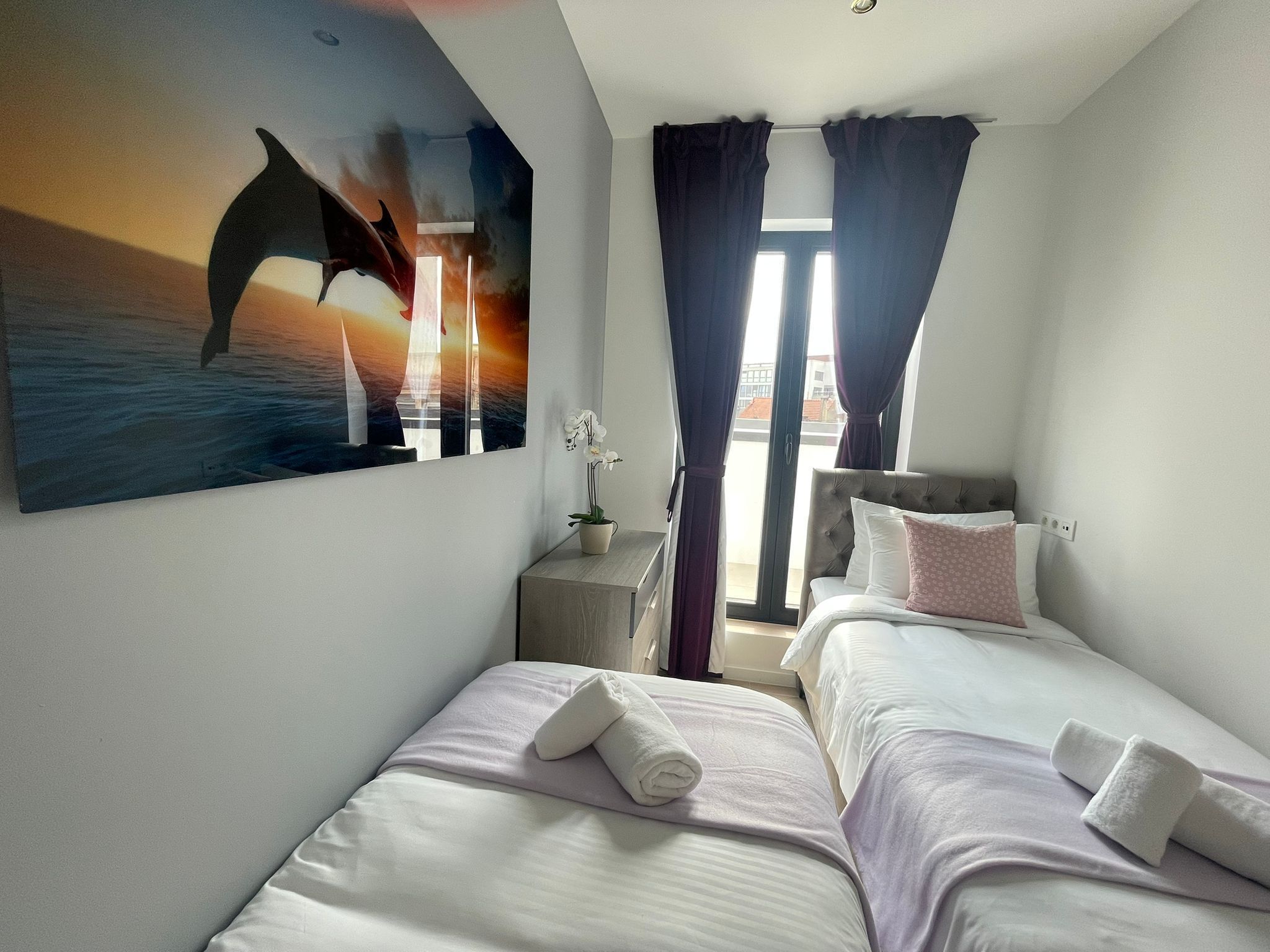 Habitación de hotel con dos camas, un cuadro de delfín, una ventana con cortinas y vistas a la ciudad.
