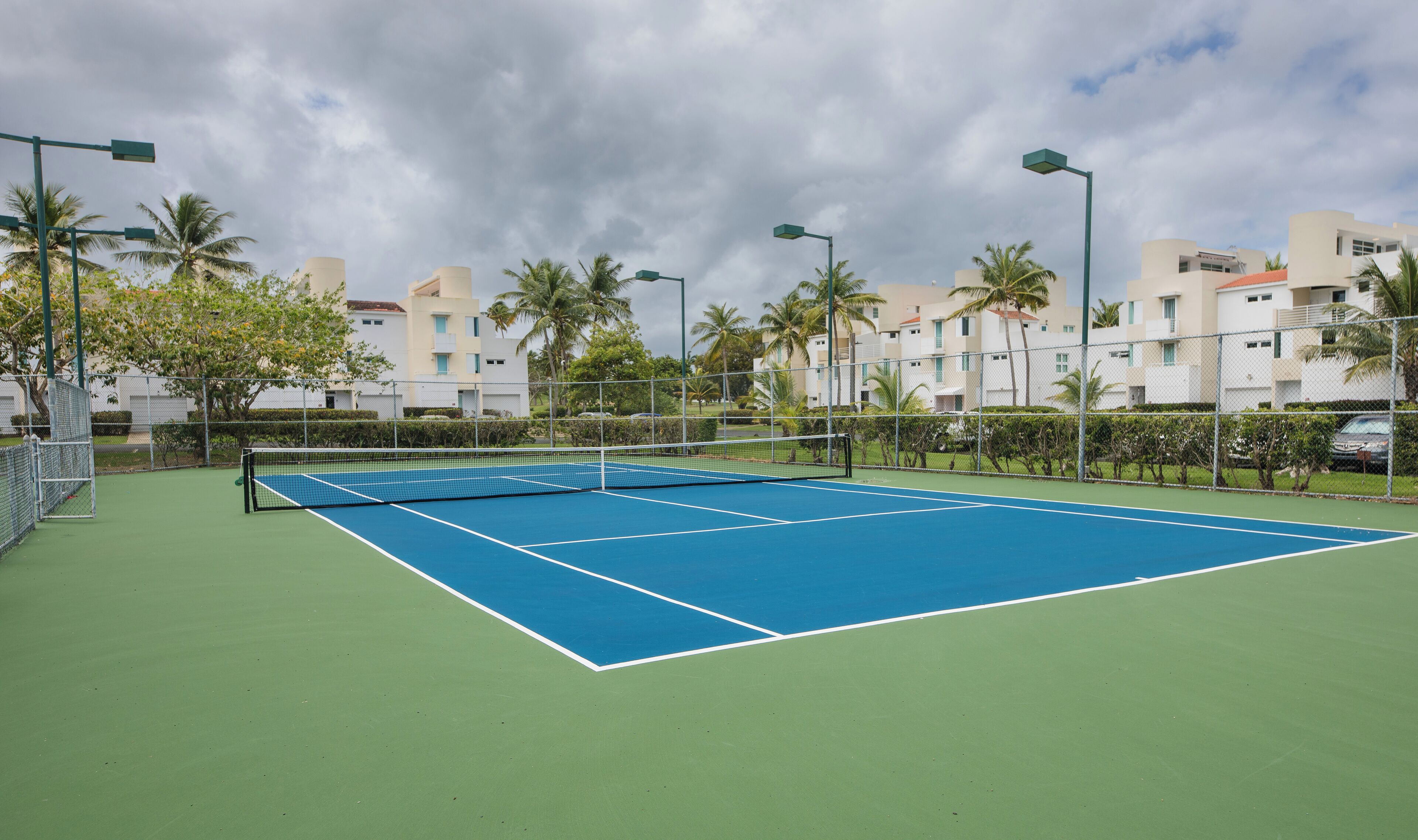 Una cancha de tenis con red, rodeada de una cerca, palmeras y edificios al fondo.
