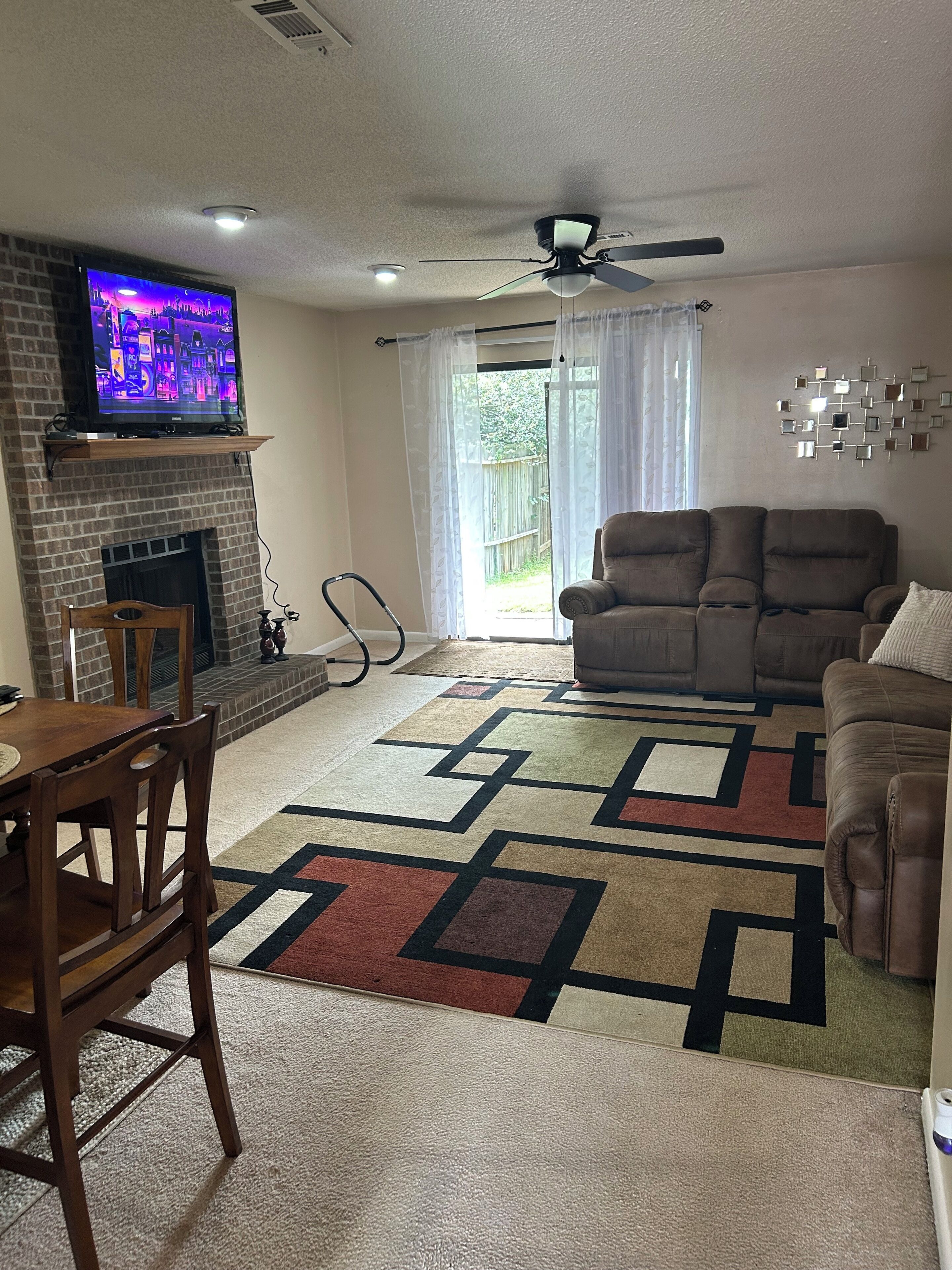 A living room with a fireplace, a television mounted above it, a ceiling fan, and a large window with curtains.