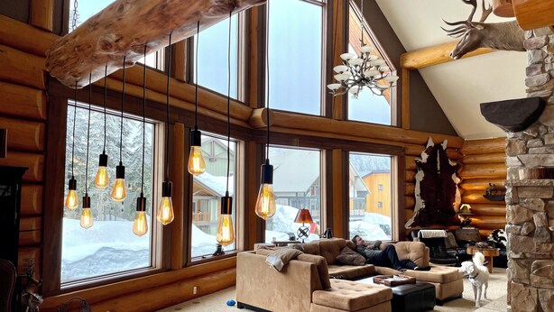 A log cabin interior with large windows, a stone fireplace, and a hanging light fixture.