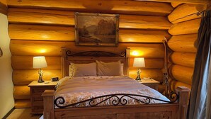 A wooden log cabin bedroom with a bed, two bedside tables with lamps, and a painting on the wall.