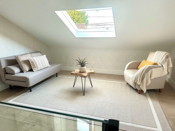 Une chambre de style loft moderne, dotée d’une lucarne, d’un canapé gris, d’un fauteuil blanc et d’une petite table en bois sur laquelle est posée une plante.