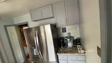 Una cocina con gabinetes de gris claro, un refrigerador de acero inoxidable y una estación de café con diversas máquinas de café y un letrero que dice 'Coffee'.