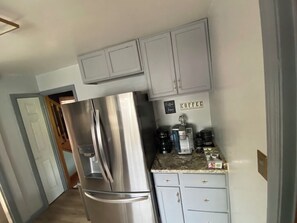 Eine Küche mit hellgrauen Schränken, einem Edelstahlkühlschrank und einer Kaffeestation mit verschiedenen Kaffeemaschinen und einem Schild mit der Aufschrift 'Coffee'.