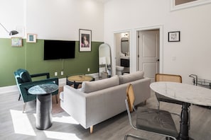 A modern living room with a grey sofa, a round coffee table, a TV, and a dining area with a white table and chairs. - Sosuite | 1BR Apt w W/D, Desk Alcove (Philadelphia)