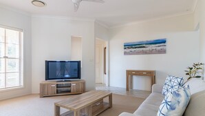Ein Wohnzimmer mit einem Flachbildfernseher auf einem Holzbodenständer, einer Couch, einem Couchtisch und einem Bild mit einer Strandansicht an der Wand.