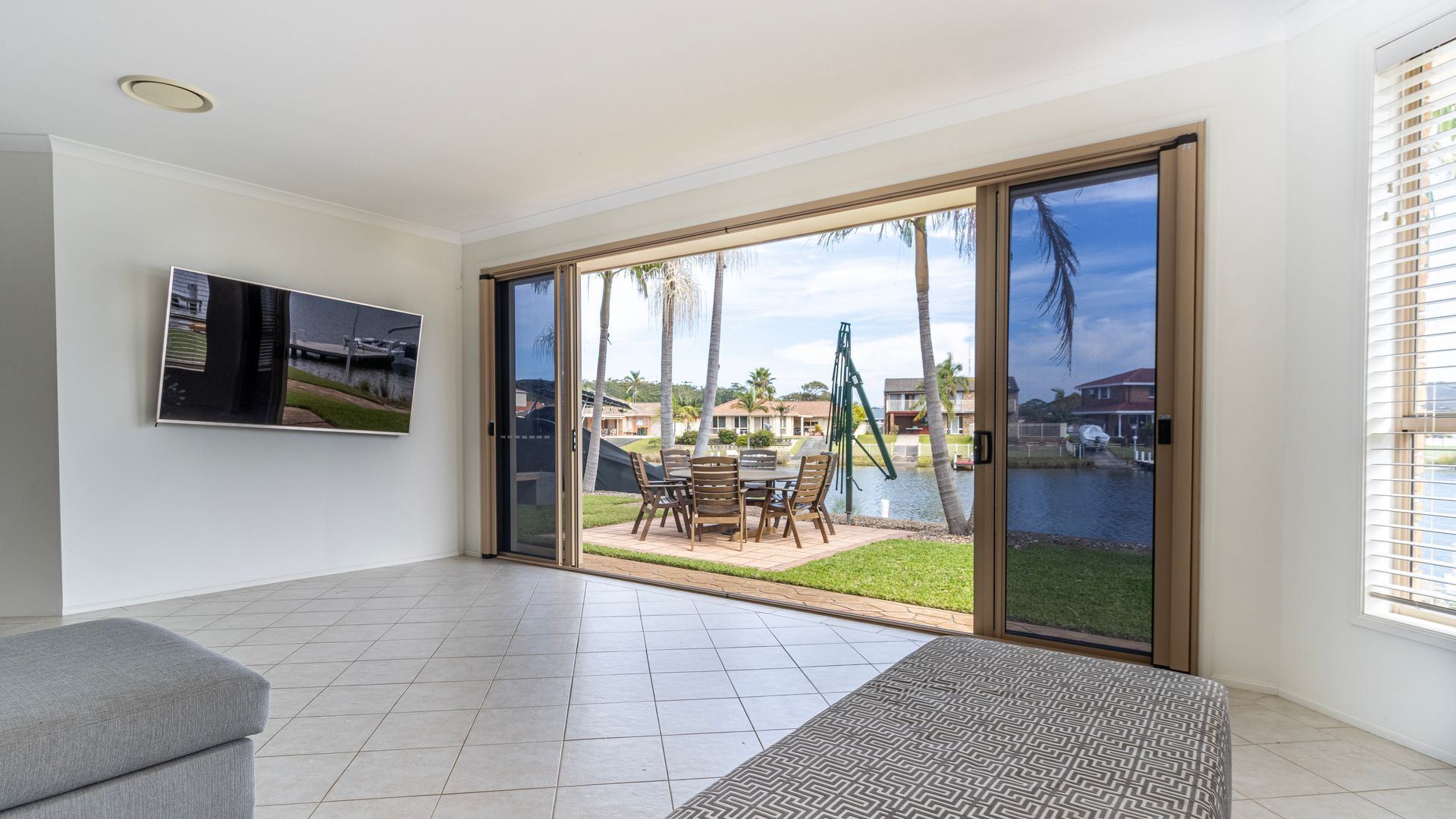 A spacious living area with a large sliding glass door leading to a poolside patio, a flat-screen TV mounted on the wall, and a grey sofa.