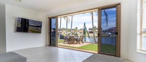A spacious living area with a large sliding glass door leading to a poolside patio, a flat-screen TV mounted on the wall, and a grey sofa.