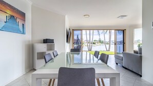 Ein modernes Wohnzimmer mit einem Glastisch, grauen Sofas und Blick auf eine Wohngegend mit Palmen und einem Steg.