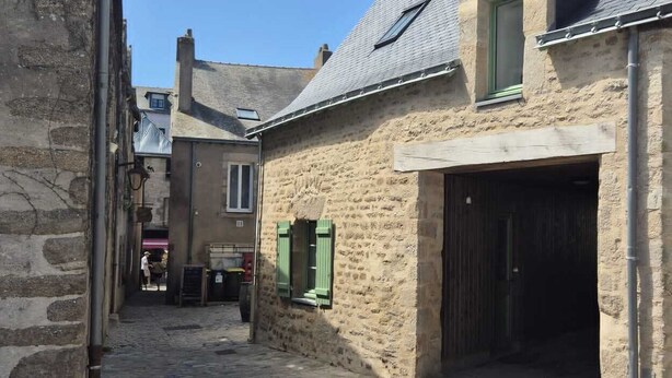Une ruelle étroite pavée de pierres, bordée de bâtiments en pierre, avec une porte de garage bien visible et un ciel bleu dégagé.