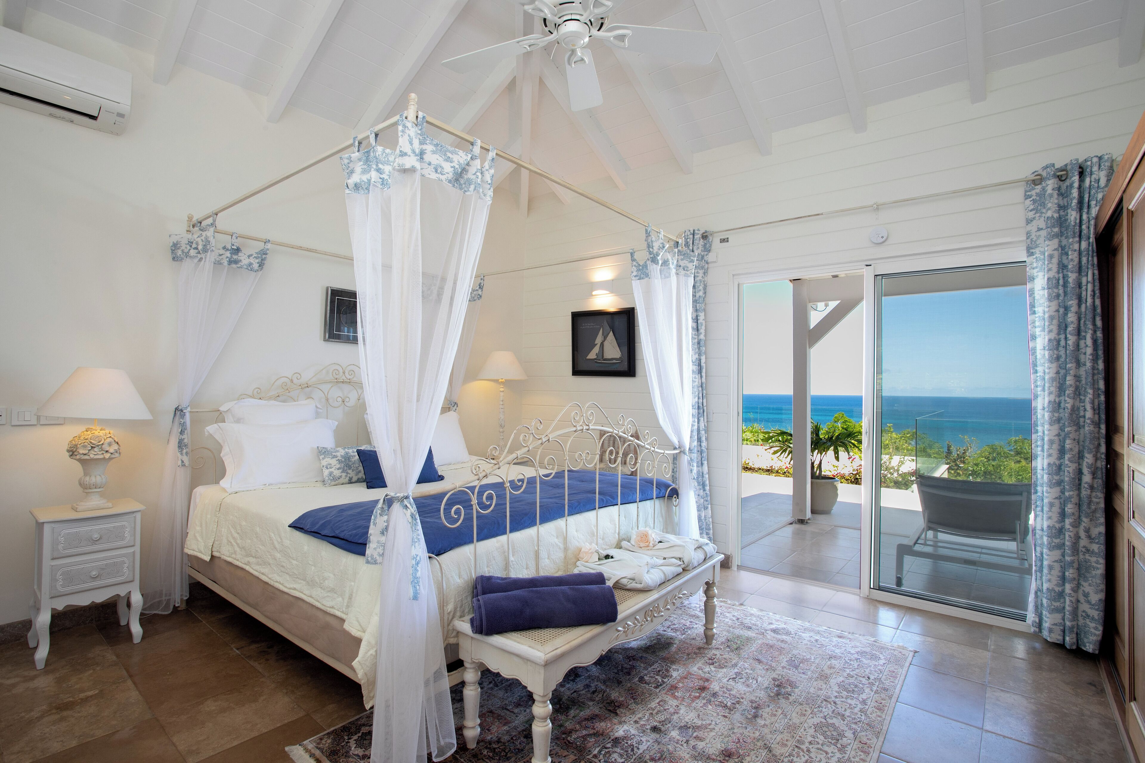 A bedroom with a four-poster bed, a nightstand with a lamp, and a view of the ocean through a sliding glass door.