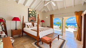 A bedroom with a four-poster bed, a wooden nightstand, a red lamp, and a view of the ocean through a glass door.